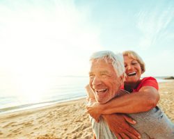 A senior couple seen having fun at the beach on a nice day. The woman is on her husband's back, with her arms crossing over the front of his chest. The sun is glistening on the ocean water.